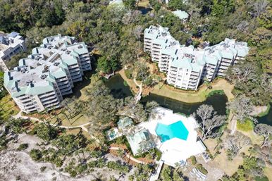 Aerial view of a beachside condominium complex with pale blue roofs, winding paths, lagoon and wooden bridge, and a bright turquoise resort pool near sand dunes and oak trees.