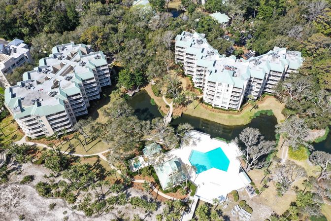 Aerial view of a beachside condominium complex with pale blue roofs, winding paths, lagoon and wooden bridge, and a bright turquoise resort pool near sand dunes and oak trees.