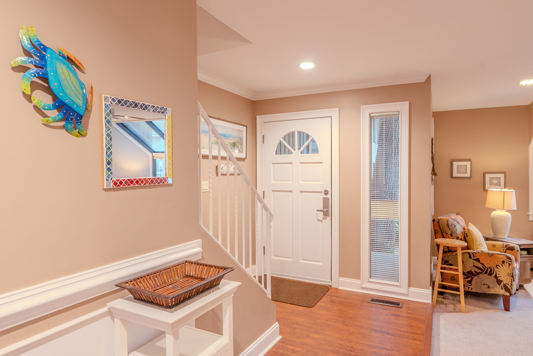 Coastal-inspired foyer with a white front door and staircase, colorful blue crab wall art and mosaic mirror above a white console with wicker tray, opening to a cozy living area with floral armchair and lamp.