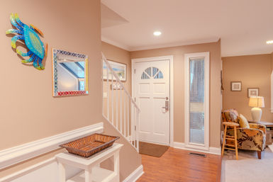 Coastal-inspired foyer with a white front door and staircase, colorful blue crab wall art and mosaic mirror above a white console with wicker tray, opening to a cozy living area with floral armchair and lamp.