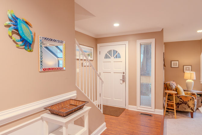 Coastal-inspired foyer with a white front door and staircase, colorful blue crab wall art and mosaic mirror above a white console with wicker tray, opening to a cozy living area with floral armchair and lamp.