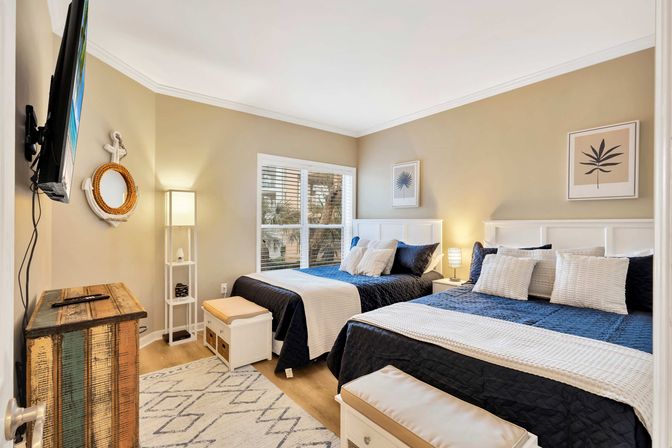 Cozy coastal guest bedroom with two navy-and-white beds, palm-tree view from window, rustic wood chest and soft bedside lighting