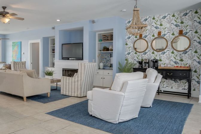 Bright coastal-inspired living room with blue walls and rugs, white upholstered armchairs and beige sofa around a fireplace and wall-mounted TV, striped accent chairs, bird-and-botanical wallpaper with three round mirrors, black console coffee station and beaded chandelier.