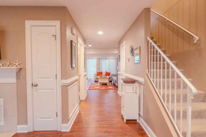 Bright modern hallway interior with hardwood floors leading to a cozy living room with red accent rug and pillows, sliding glass door, and a carpeted staircase with white railing.