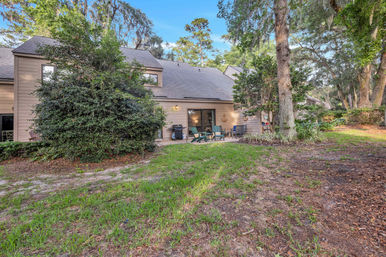 Cozy backyard patio of a two-story house framed by oak trees with Spanish moss, featuring a sliding glass door, Adirondack chairs, a grill, and a grassy yard.