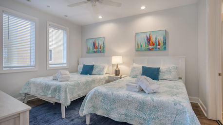 Bright beach-style bedroom with two beds in seafoam quilts, blue accent pillows, folded towels, nightstand lamp, colorful sailboat paintings, three windows and ceiling fan