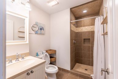 Bright modern bathroom with white vanity and mirror, chrome fixtures, toilet with wicker shelving, and a walk-in beige tiled shower with a white curtain