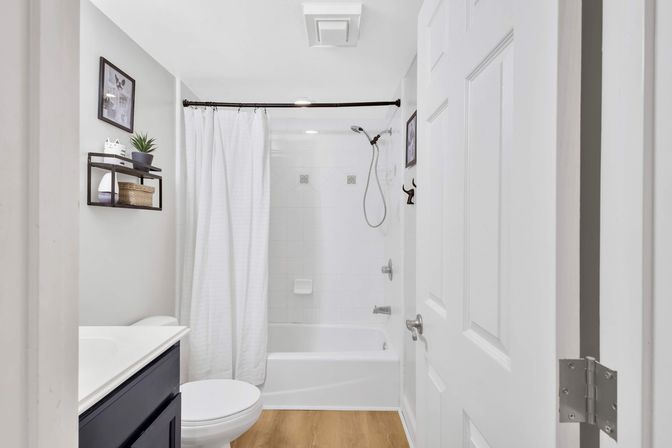 Bright contemporary small bathroom with white tiled tub-shower and curtain, handheld showerhead, dark vanity, wood-look floor, and wall shelf with plant and basket