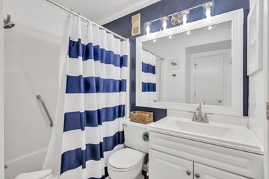 Small coastal-style bathroom interior with navy-and-white striped shower curtain, white vanity and sink, large framed mirror with vanity lights, toilet with wicker basket and navy accent wall