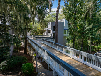 Sun-dappled elevated wooden boardwalk winding through live oak trees draped with Spanish moss, connecting multi-level coastal residences amid lush, shaded landscaping.