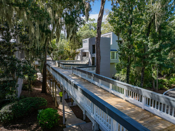 Sun-dappled elevated wooden boardwalk winding through live oak trees draped with Spanish moss, connecting multi-level coastal residences amid lush, shaded landscaping.
