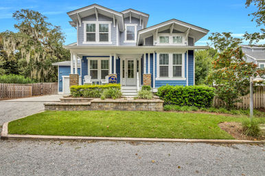 Blue Craftsman-style two-story house with white trim and covered front porch featuring Adirondack chairs, raised brick planter and manicured green lawn, driveway and wooden fence, trees draped in Spanish moss.