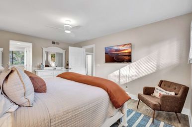 Bright coastal-style bedroom with white quilted bed, rust-orange throw, white dresser and mirror, wall-mounted TV showing a beach sunset, brown accent chair and sunlit blinds.