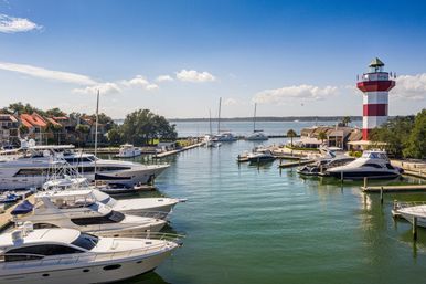 Coastal marina with luxury yachts docked along a calm channel, red-and-white striped lighthouse on the right, waterfront homes and sailboats under a sunny blue sky
