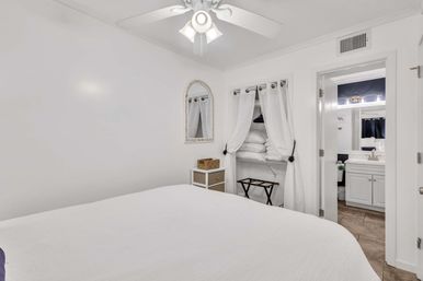 Bright white bedroom with ceiling fan, neatly made bed, open curtained closet stacked with linens, small dresser and doorway to a white en-suite bathroom.