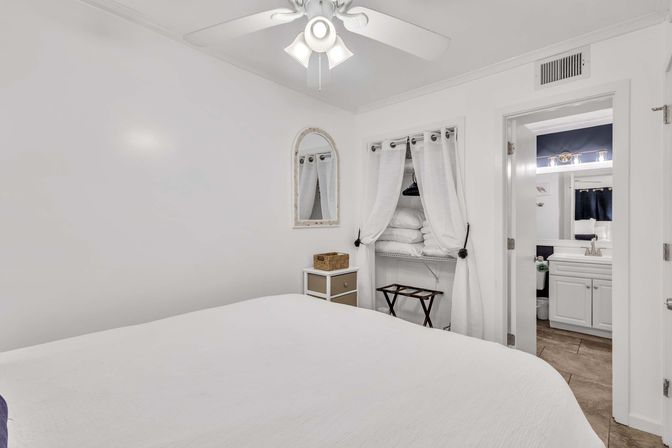Bright white bedroom with ceiling fan, neatly made bed, open curtained closet stacked with linens, small dresser and doorway to a white en-suite bathroom.