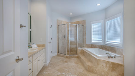 Bright modern master bathroom with beige marble tile floors and surround, glass-enclosed corner shower, raised soaking tub with steps and chrome faucet, white vanity with sink, and two windows with blinds.