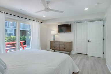 Bright modern bedroom retreat with white quilted bed, ceiling fan, wall-mounted TV above a mid-century dresser, and sliding glass doors opening to a patio with a red picnic table and lush greenery.