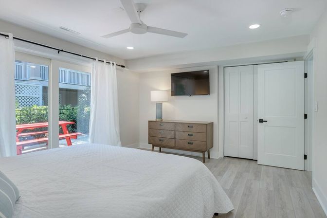 Bright modern bedroom retreat with white quilted bed, ceiling fan, wall-mounted TV above a mid-century dresser, and sliding glass doors opening to a patio with a red picnic table and lush greenery.