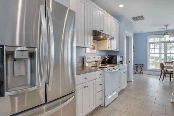 Bright coastal-style kitchen with stainless steel French-door refrigerator, white shaker cabinets, granite countertops, tile floor, electric range and microwave, and a dining nook by a large window