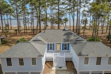 Elevated coastal beach house with gray shingled roof, blue shutters and white front stairs, nestled among tall pine trees with the ocean visible in the background.