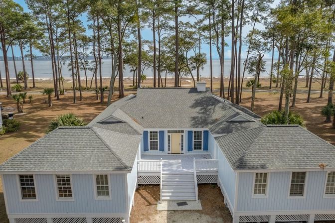 Elevated coastal beach house with gray shingled roof, blue shutters and white front stairs, nestled among tall pine trees with the ocean visible in the background.