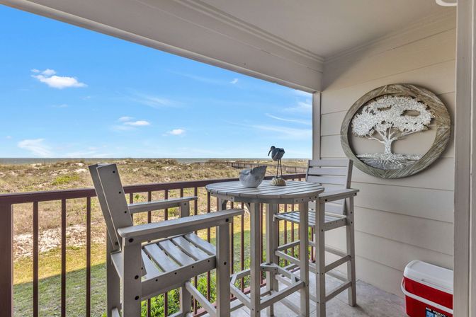 Coastal balcony with bar-height bistro table and chairs overlooking sand dunes and ocean, with a whimsical bird sculpture on the table and round tree wall art on the wall.