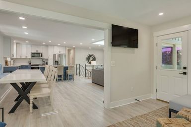 Bright open-concept modern farmhouse kitchen and dining area with white cabinets, navy island, light wood floors, recessed lighting, wall-mounted TV and glass-paneled entry door.