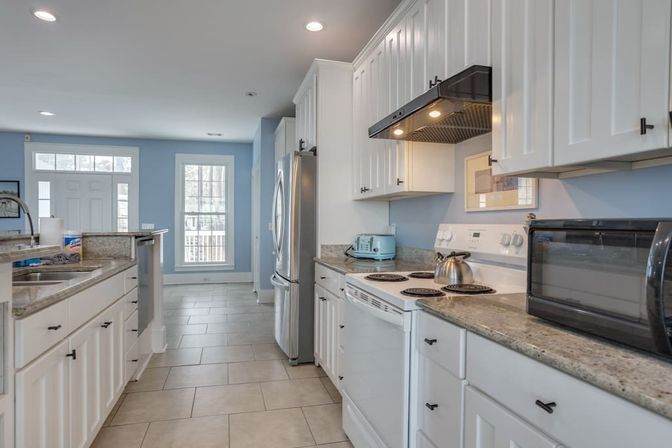 Bright modern galley kitchen with white shaker cabinets, granite countertops, stainless refrigerator, electric stove with kettle, blue walls, tile floor and countertop toaster and microwave
