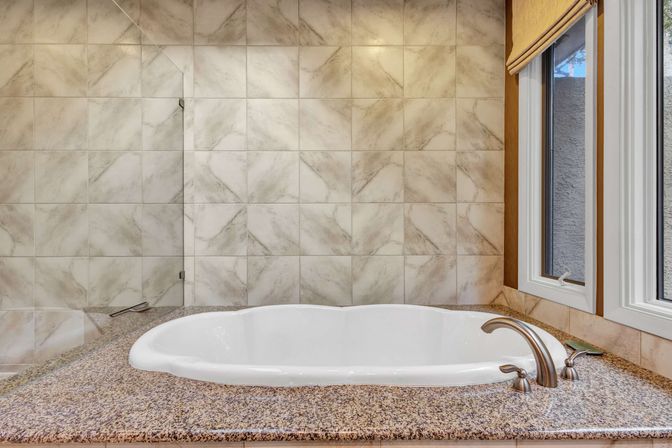 Spa-like master bathroom with white built-in soaking tub set in speckled granite surround, brushed nickel faucet, marble-look tiled wall and window with Roman shade