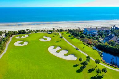 Aerial view of an oceanfront golf course with vivid green fairways, curvy sand bunkers, palm-lined cart path, seaside homes and a sandy beach meeting turquoise ocean.