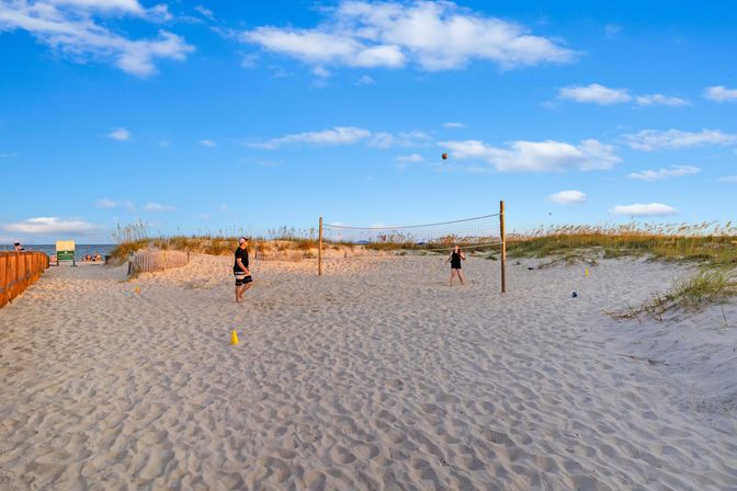 Two people playing beach volleyball on a sandy coastal court with grassy dunes, a wooden walkway and ocean visible in the background under a bright blue sky.