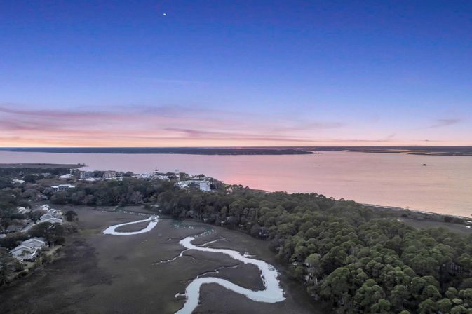 Aerial view of a coastal estuary at sunset — a winding tidal creek snaking through salt marsh, dense pine forest, and a shoreline town along a calm pink-blue bay.