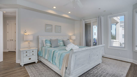 Sunlit coastal-style master bedroom with a white distressed sleigh bed, teal-and-aqua striped bedding, matching nightstands and lamps, sliding glass door to a balcony, hardwood floors and area rug.