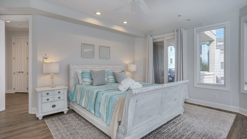 Sunlit coastal-style master bedroom with a white distressed sleigh bed, teal-and-aqua striped bedding, matching nightstands and lamps, sliding glass door to a balcony, hardwood floors and area rug.