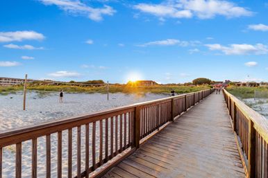 Wooden boardwalk leading across sandy beach to coastal dunes and a beach volleyball court at golden sunset under a bright blue sky with scattered clouds.
