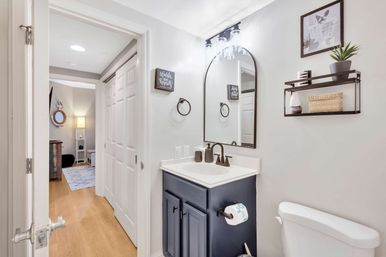 Bright modern residential bathroom interior featuring a navy vanity with white countertop, arched mirror and matte-black fixtures, a wall shelf with plant and wicker basket, toilet, and an open doorway to a wood‑floored hallway and bedroom.