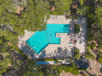 Aerial view of a sun-dappled turquoise outdoor pool with loungers and a few swimmers on a paved deck, nestled among lush trees and landscaped greenery for a relaxed vacation vibe.
