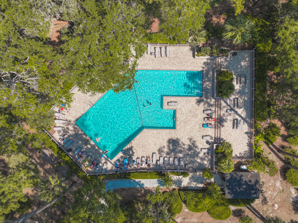 Aerial view of a sun-dappled turquoise outdoor pool with loungers and a few swimmers on a paved deck, nestled among lush trees and landscaped greenery for a relaxed vacation vibe.
