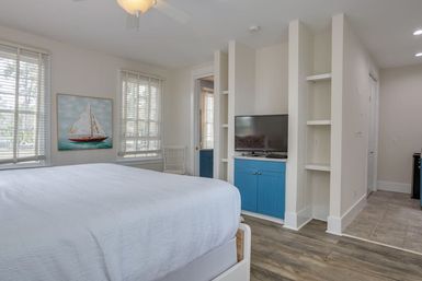 Bright coastal-inspired bedroom with white bedding, sailboat painting, twin windows with blinds, ceiling fan, built-in shelving and blue media cabinet with flat-screen TV over wood-look floors
