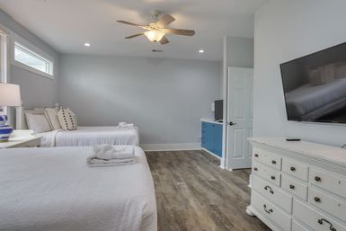 Bright coastal-style bedroom with two white-made beds, folded towels, ceiling fan, blue cabinet, wall-mounted TV and wood-look flooring