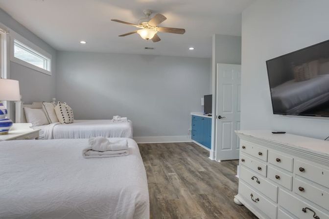 Bright coastal-style bedroom with two white-made beds, folded towels, ceiling fan, blue cabinet, wall-mounted TV and wood-look flooring