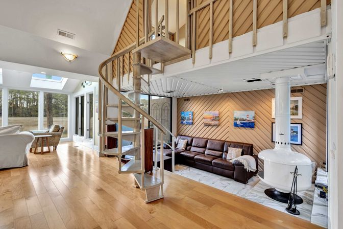 Sunlit coastal living area with a spiral metal staircase to a loft, brown leather sectional against a chevron wood accent wall, white freestanding stove, and floor-to-ceiling windows overlooking marshland.