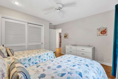 Bright coastal-themed guest bedroom with two twin beds in seashell-patterned quilts, white dresser, ceiling fan, sliding louvered closet doors, hardwood floor and teal curtains