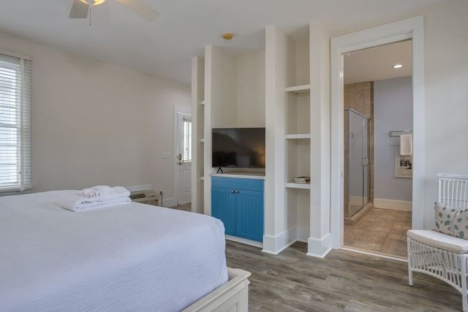 Bright coastal-style bedroom with white bed and folded towels, built-in shelving and blue media cabinet with flatscreen TV, wood-look flooring, wicker chair, ceiling fan, and doorway to tiled en-suite bathroom with glass shower