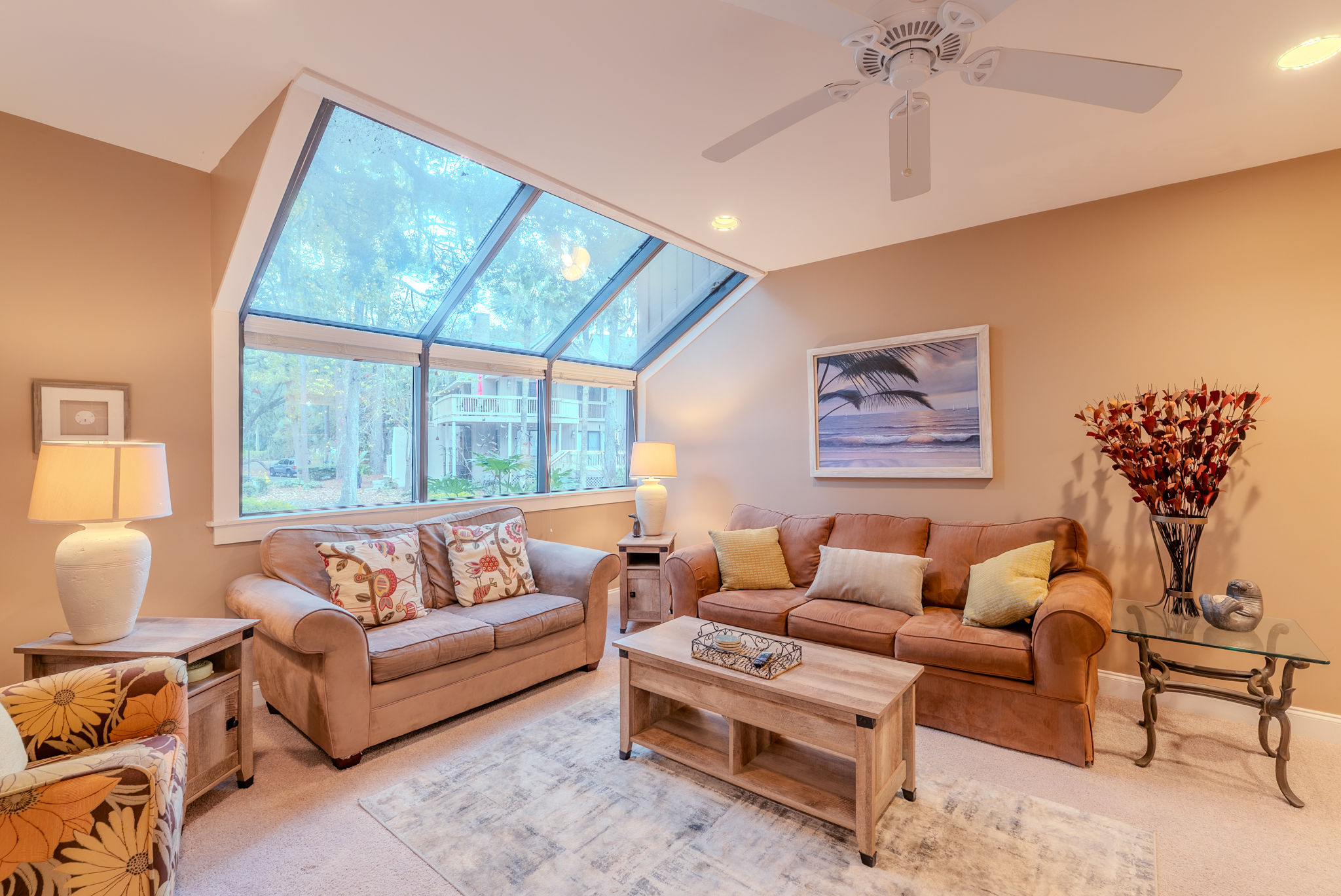 Sunlit cozy living room with large angled skylight windows overlooking trees, two beige sofas, wooden coffee table, coastal wall art, table lamps and decorative vase of dried flowers