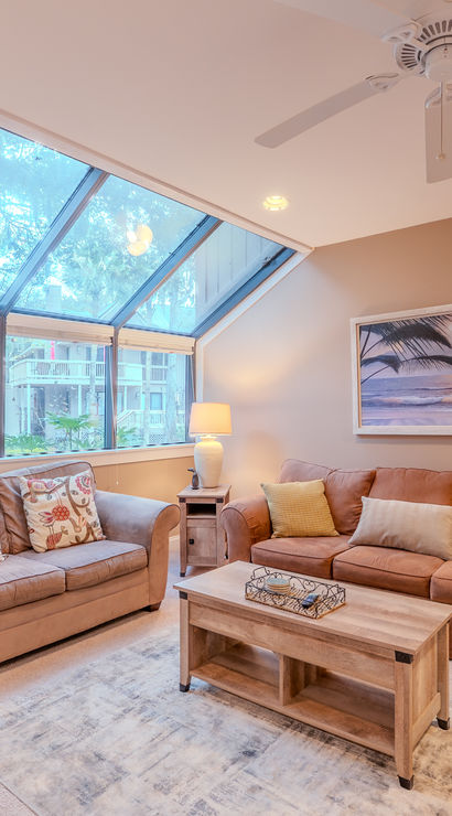 Sunlit cozy living room with large angled skylight windows overlooking trees, two beige sofas, wooden coffee table, coastal wall art, table lamps and decorative vase of dried flowers