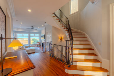Sunlit coastal interior with curved wooden staircase and wrought-iron railing, hardwood floors, large windows framing ocean and marsh views, neutral sectional and lamp.