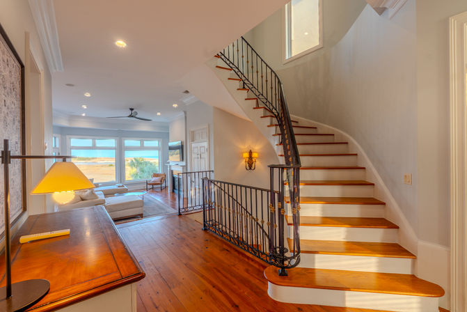 Sunlit coastal interior with curved wooden staircase and wrought-iron railing, hardwood floors, large windows framing ocean and marsh views, neutral sectional and lamp.