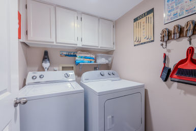 Cozy home laundry room with white top‑load washer and dryer under white cabinets, shelf with folded colorful towels, wall hooks holding a broom and red dustpan, and a golf poster on beige walls.